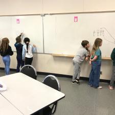 A group of students working on large whiteboards on the wall, completing math equations