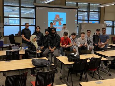 Group of 12 students with 1 male teacher sits on desks to pose for picture
