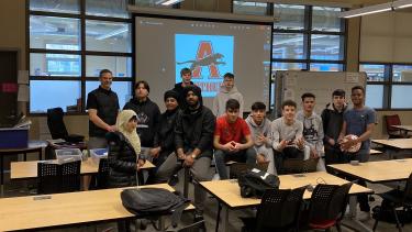 Group of 12 students with 1 male teacher sits on desks to pose for picture