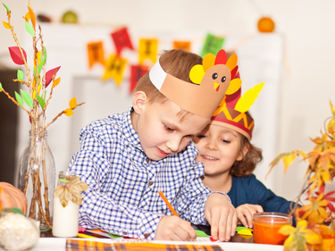 Children wearing handmade Thanksgiving-themed paper hats while creating autumn crafts at a table decorated with fall leaves and seasonal decorations.